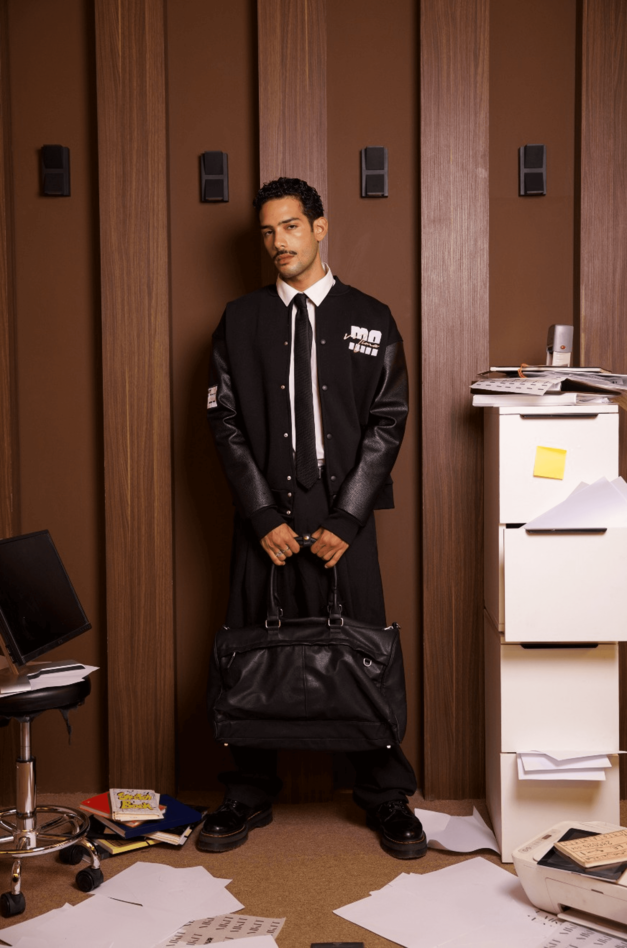 Senior in a black varsity jacket holding a duffel bag in a cluttered office